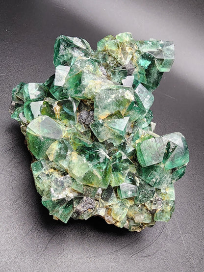 A vibrant green crystal cluster from The Crystalary, featuring well-defined cubic formations of fluorite, rests on a dark gray surface. Sourced from the Diana Marie Mine in Weardale, County Durham, England, the natural translucent green hues of the Fluorite w/ Galena—enhanced with hints of clear and darker shades—exhibit an intricate geometric structure beautifully accented by embedded galena crystals.