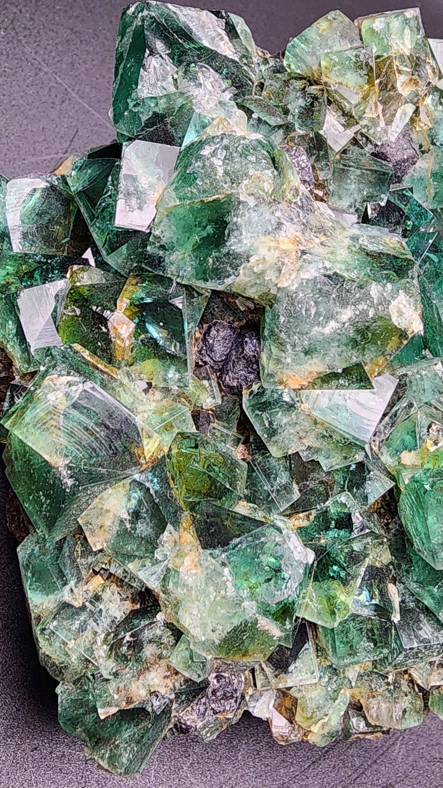 A stunning cluster of vibrant green fluorite crystals interspersed with galena crystals from The Crystalary's Fluorite w/ Galena- Heavy Metal Pocket, sourced from the Diana Marie Mine in Weardale, County Durham, England, is displayed on a dark surface. The angular formations vary in size and are closely packed together, showcasing their natural geometric arrangements with some translucent and white regions.