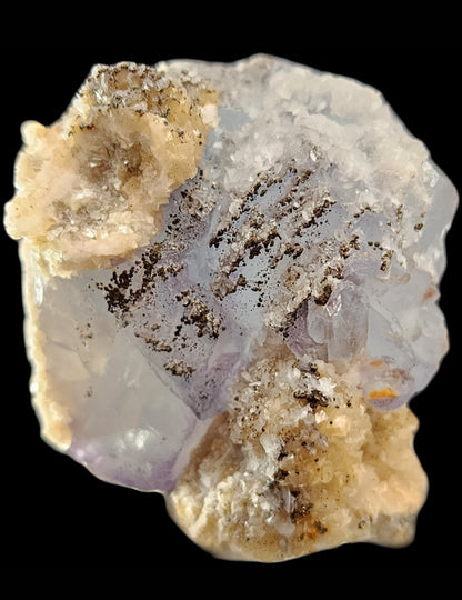 Close-up of a rough, translucent Fluorite crystal with a light purple hue and icy blue undertones from The Crystalary's Yaogangxian collection, featuring white and pale beige mineral deposits. Black specks and thin streaks are scattered across its surface, highlighting the intricate texture of the Fluorite against a black background.