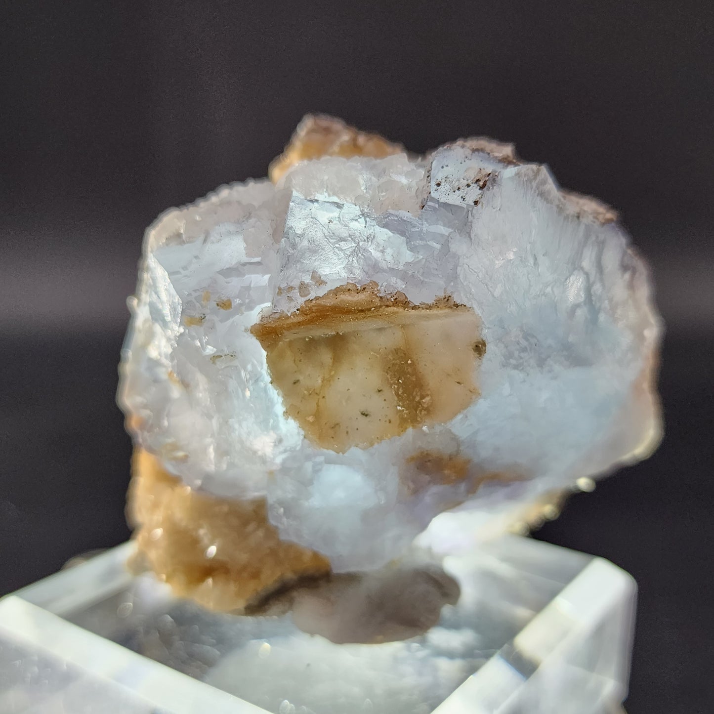 A close-up image of a translucent Fluorite specimen from Yaogangxian, offered by The Crystalary, with an icy blue hue, displaying a rough, crystal-like texture. The mineral is mounted on a clear, square base, and the dark background highlights its intricate details and subtle color variations.