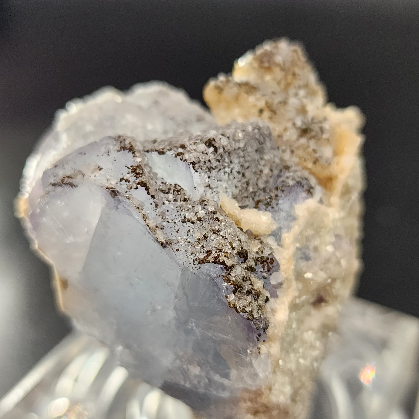 A close-up photograph showcases an iridescent Fluorite, Yaogangxian specimen from The Crystalary, featuring rough, textured surfaces. The mineral exhibits a blend of icy blue, white, and brown hues and is set against a dark, blurred background. The crystal structure appears intricate and multifaceted.