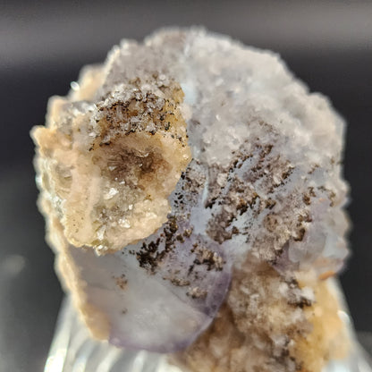 A detailed image showcasing a Fluorite, Yaogangxian from The Crystalary reveals its rough, crystalline texture. The mineral's surface features a blend of white, beige, and subtle purple hues interspersed with black specks. Among the crystals lies an icy blue Fluorite specimen against a dark, softly blurred background.