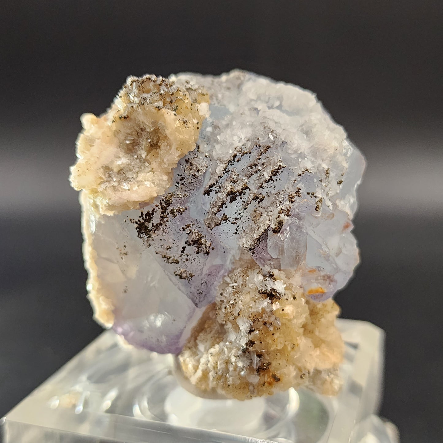 A close-up image of The Crystalary's Fluorite, Yaogangxian specimen, showcasing its pale purple cubic formations partially draped in beige and white crystalline deposits. The piece is elegantly displayed on a transparent stand against a gradient dark background, revealing subtle hints of icy blue hues.