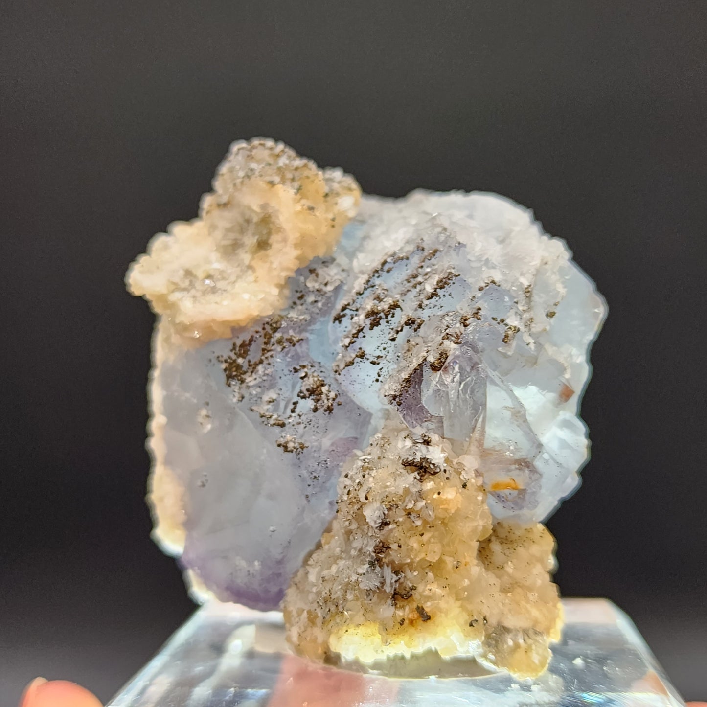 A close-up of a Yaogangxian Fluorite specimen from The Crystalary displayed on a clear stand. The mineral, with its icy blue hues and rough texture, features patches of white and brown crystallization. Set against a dark background, the intricate details of this beautiful Fluorite piece are highlighted brilliantly.