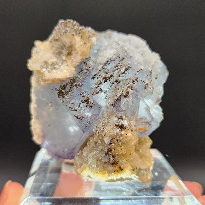 A close-up photograph showcases the Yaogangxian Fluorite, a translucent, icy blue crystalline mineral specimen with light blue and white hues, accented by patches of dark gray inclusions. This stunning piece from The Crystalary is elegantly positioned on a clear display stand against a black background, emphasizing its intricate textures and vibrant colors.