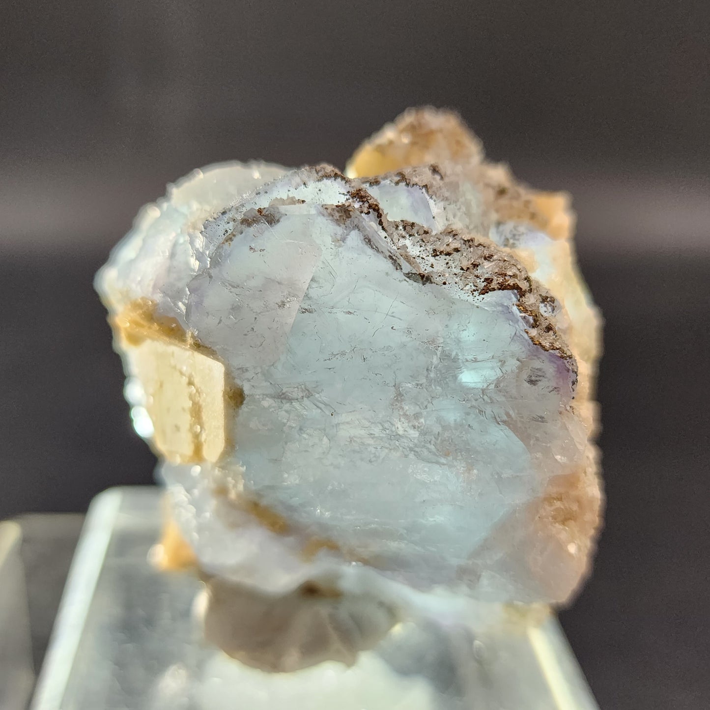 Close-up of The Crystalary's Fluorite, Yaogangxian specimen with icy blue and gray hues, elegantly positioned on a clear display stand. The mineral features rough edges with some beige inclusions and reflective surfaces. The blurred background accentuates the mineral's intricate texture and details.