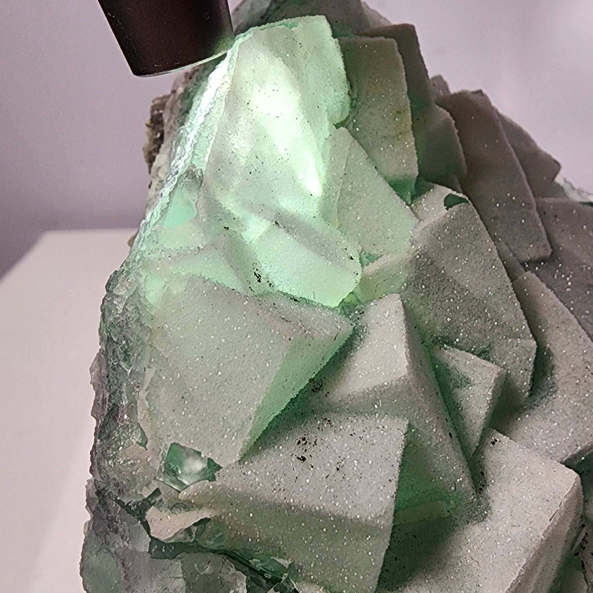 A close-up image of The Crystalary's Fluorite-Yaogangxian with Mico "Sugar" Quartz, illuminated by an ultraviolet light source from above. The large, translucent seafoam green fluorite crystal features cubic formations and a slightly rough texture, with a light-green glow emanating from the illuminated areas. This exquisite specimen weighs 248 grams.