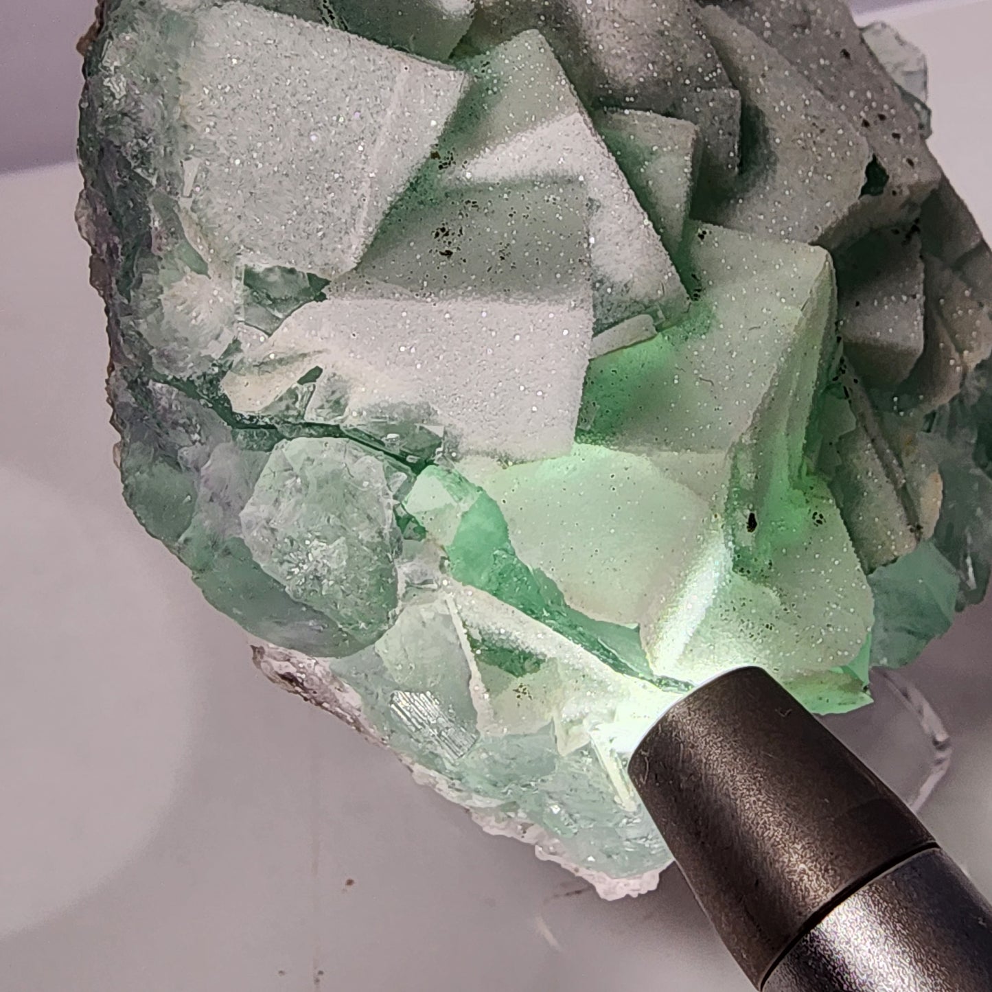 A flashlight illuminates a large, crystalline formation of seafoam green Fluorite-Yaogangxian with light green and white cubic crystals from The Crystalary. The textured surface is sparkly and intricate, with rough edges and layered geometric shapes. The sugary micro quartz enhances the clarity and color contrasts.