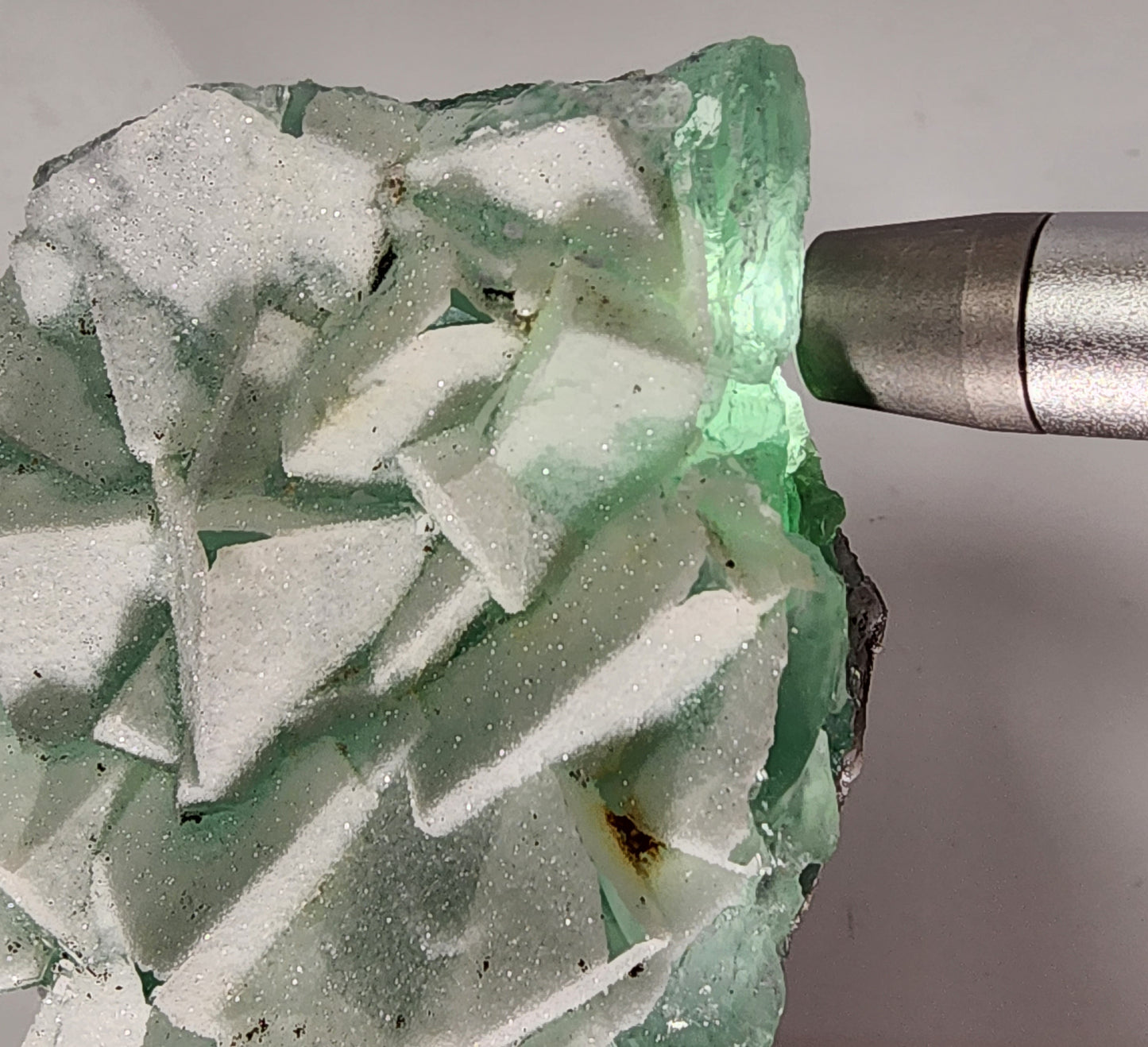 A close-up of the Fluorite-Yaogangxian with Mico "Sugar" Quartz specimen by The Crystalary, showcasing its seafoam green hue and rough, angular surface. A metal flashlight is partially visible on the right side, illuminating the 248-gram specimen and highlighting its translucent and crystalline structures.