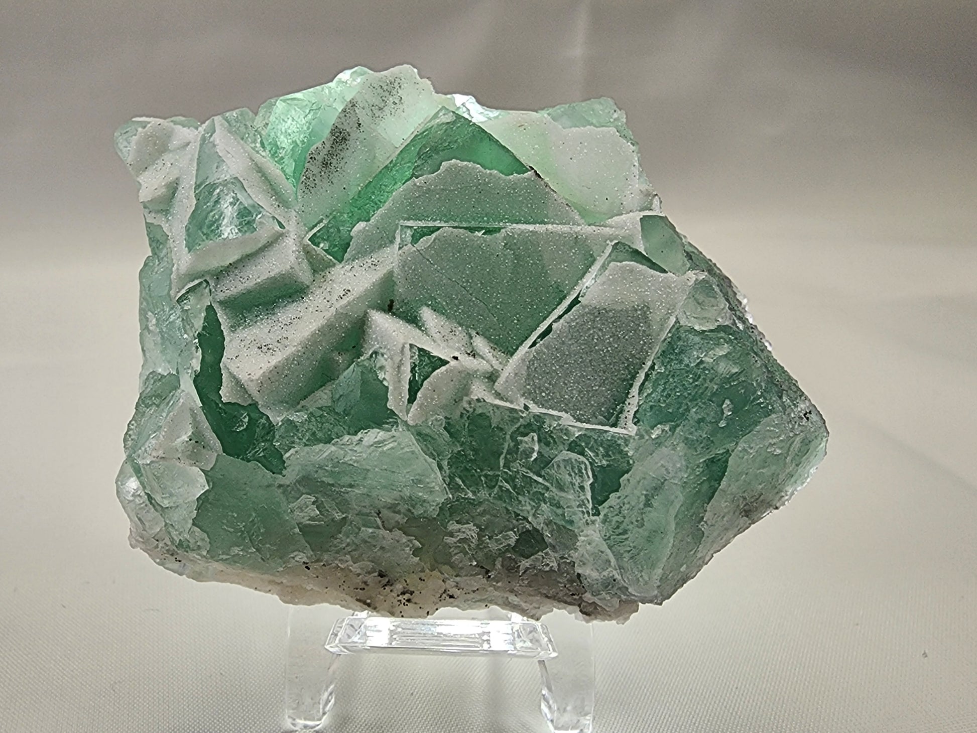 A close-up of the Fluorite-Yaogangxian with Micro "Sugar" Quartz mineral specimen by The Crystalary, displayed on a clear plastic stand. The seafoam green fluorite crystal features a rough, jagged surface interspersed with translucent and opaque areas that capture light to create an array of light and dark green hues. The background is a plain, light gray.