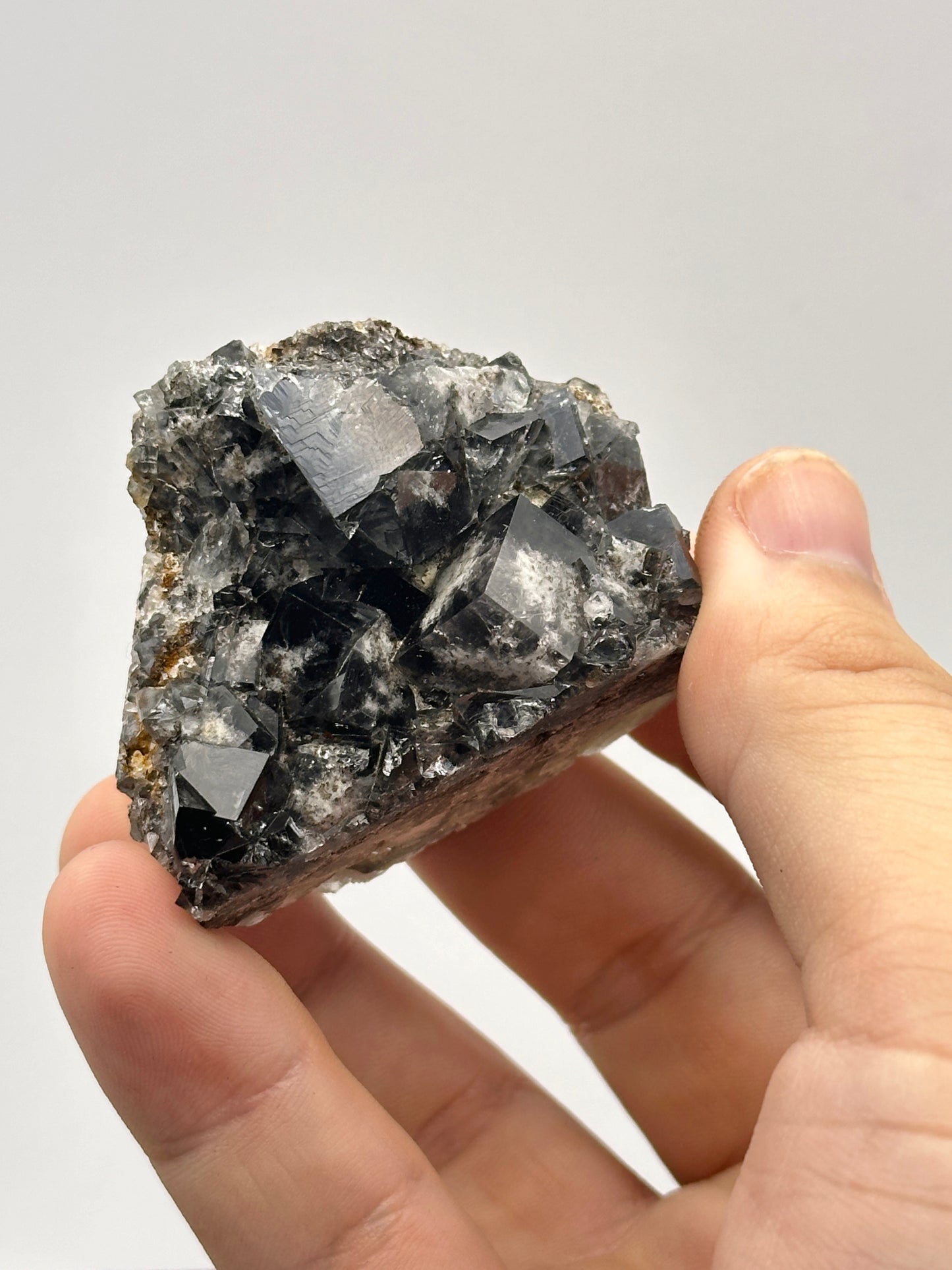 A hand holds a jagged, black Fluorite cluster from the Yum Yum Pocket at Diana Maria Mine against a plain, light background. The crystals from The Crystalary have a shiny, reflective surface with uneven, natural formations. The fingers are gently wrapped around the edges, highlighting the raw, unpolished texture of this specimen from Frosterley in Weardale, England.