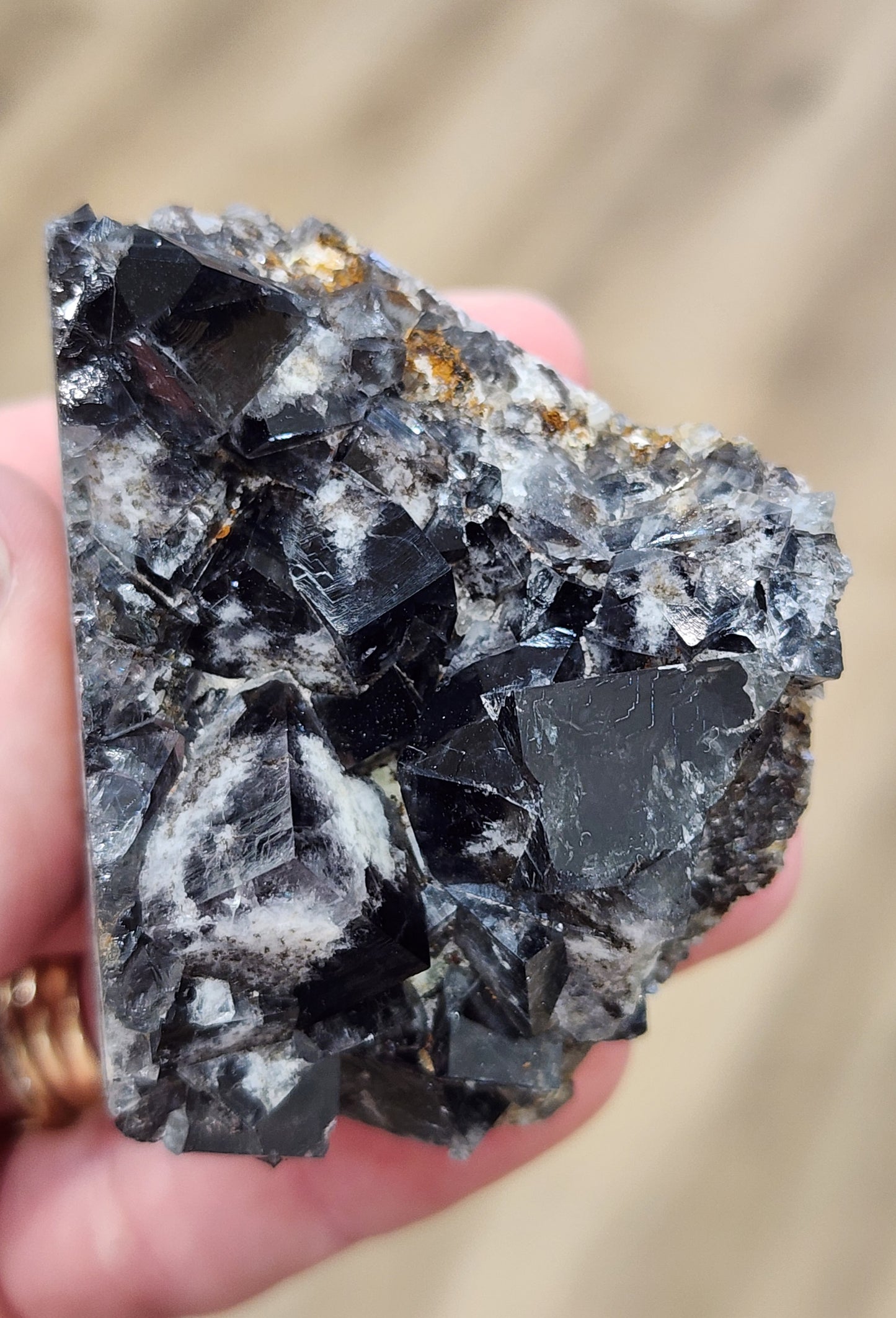 A close-up of a hand holding The Crystalary's Fluorite from the Yum Yum Pocket, Diana Maria Mine in Frosterley, Weardale, England. The mineral is mostly black with silver and gray streaks, displaying a rough, jagged texture. The background is slightly blurred, putting the focus on the intricacies of the mineral's surface.