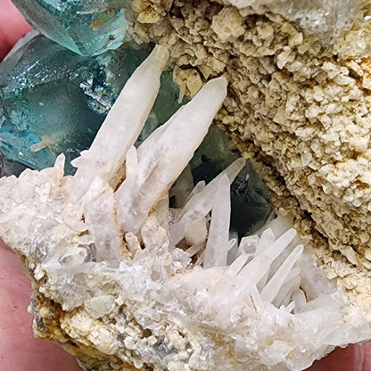 Close-up of a mineral specimen from The Crystalary, with sharp, white quartz crystals jutting out from a textured, beige matrix. In the background, cubic green fluorite crystals are densely aggregated, displaying a translucent to slightly opaque appearance—a striking association of Fluorite and Milky Quartz from the Brandberg Area in Dâures Constituency, Erongo Region, Namibia.