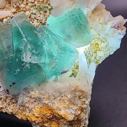 A close-up photo of a Fluorite/Milky Quartz specimen by The Crystalary features large, sea green aqua fluorite crystals surrounded by small white quartz crystals on a beige, textured rock matrix. Sourced from the Brandberg Area in the Dâures Constituency of Namibia's Erongo Region, this stunning mineral arrangement is set against a neutral dark background to highlight the vibrant colors and milky quartz association.