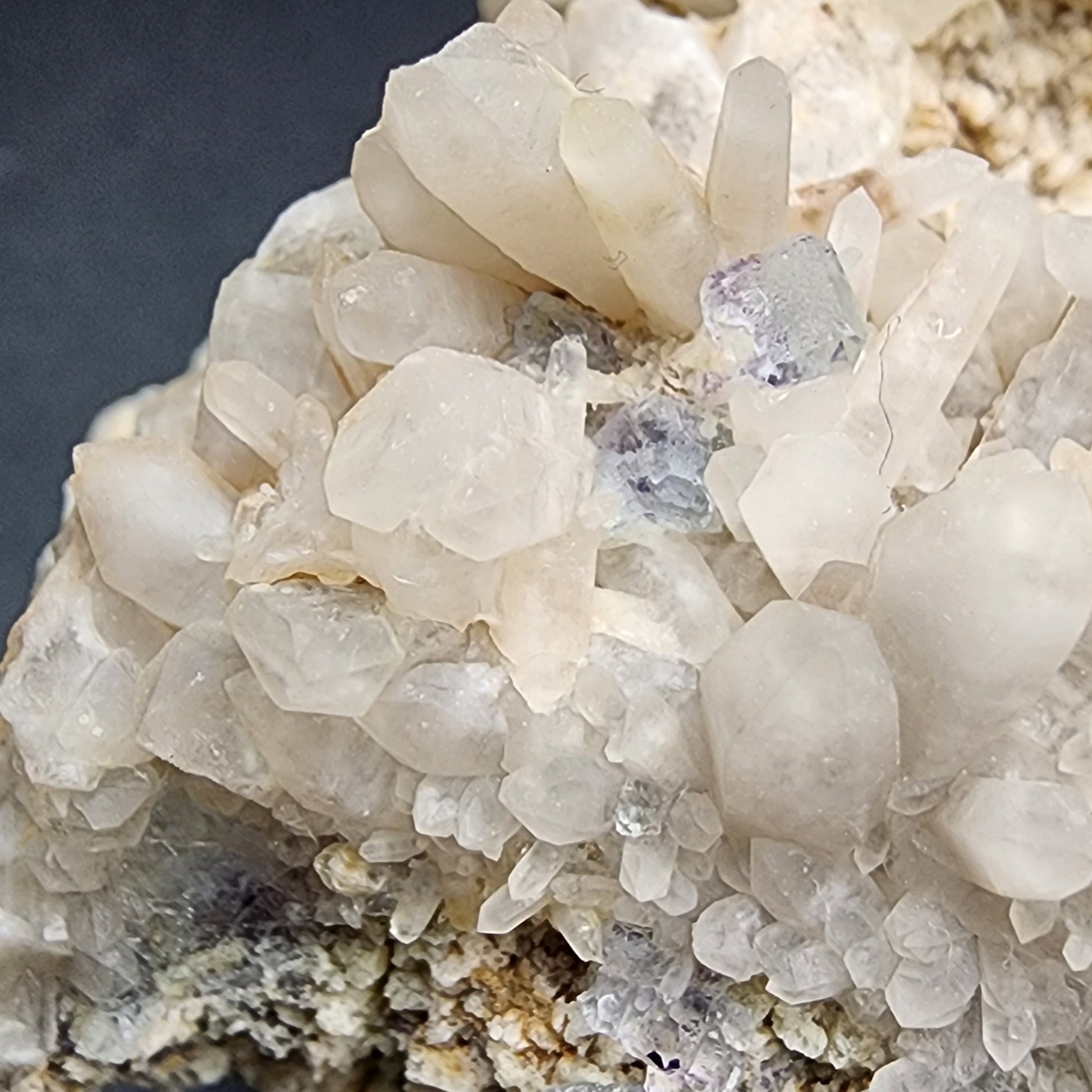 A close-up image of a cluster of Fluorite/Milky Quartz crystals from the Brandberg Area, Dâures Constituency, Erongo Region, Namibia showcases transparent sections with hints of purple hues growing from a rocky surface. The jagged, intricate texture reveals various formations subtly hinting at its milky quartz association. Available from The Crystalary.