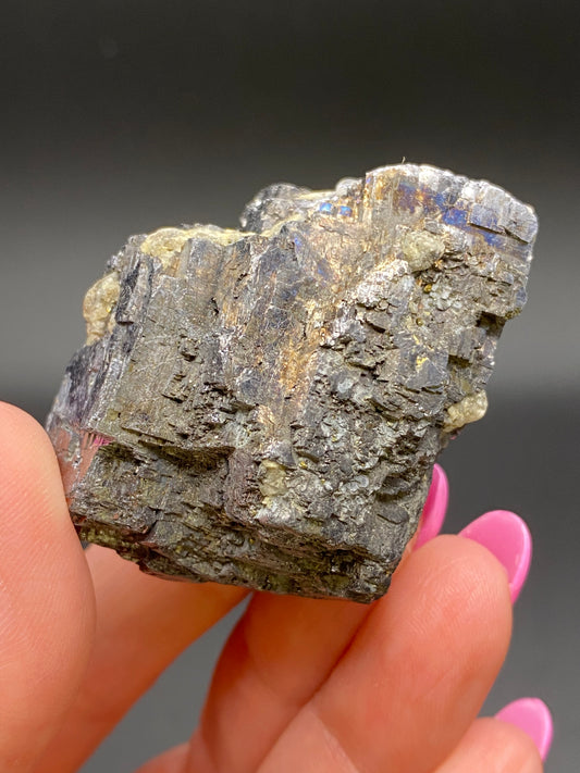 A hand with pink-painted nails holds a rough, irregularly shaped mineral rock featuring a mix of dark gray and golden areas. The Galena rock from the Sweetwater Mine in Ellington, Reynolds County, Missouri, has a textured, layered appearance with a combination of crystalline and earthy surfaces. The background is solid black. This exquisite specimen is part of The Crystalary collection.