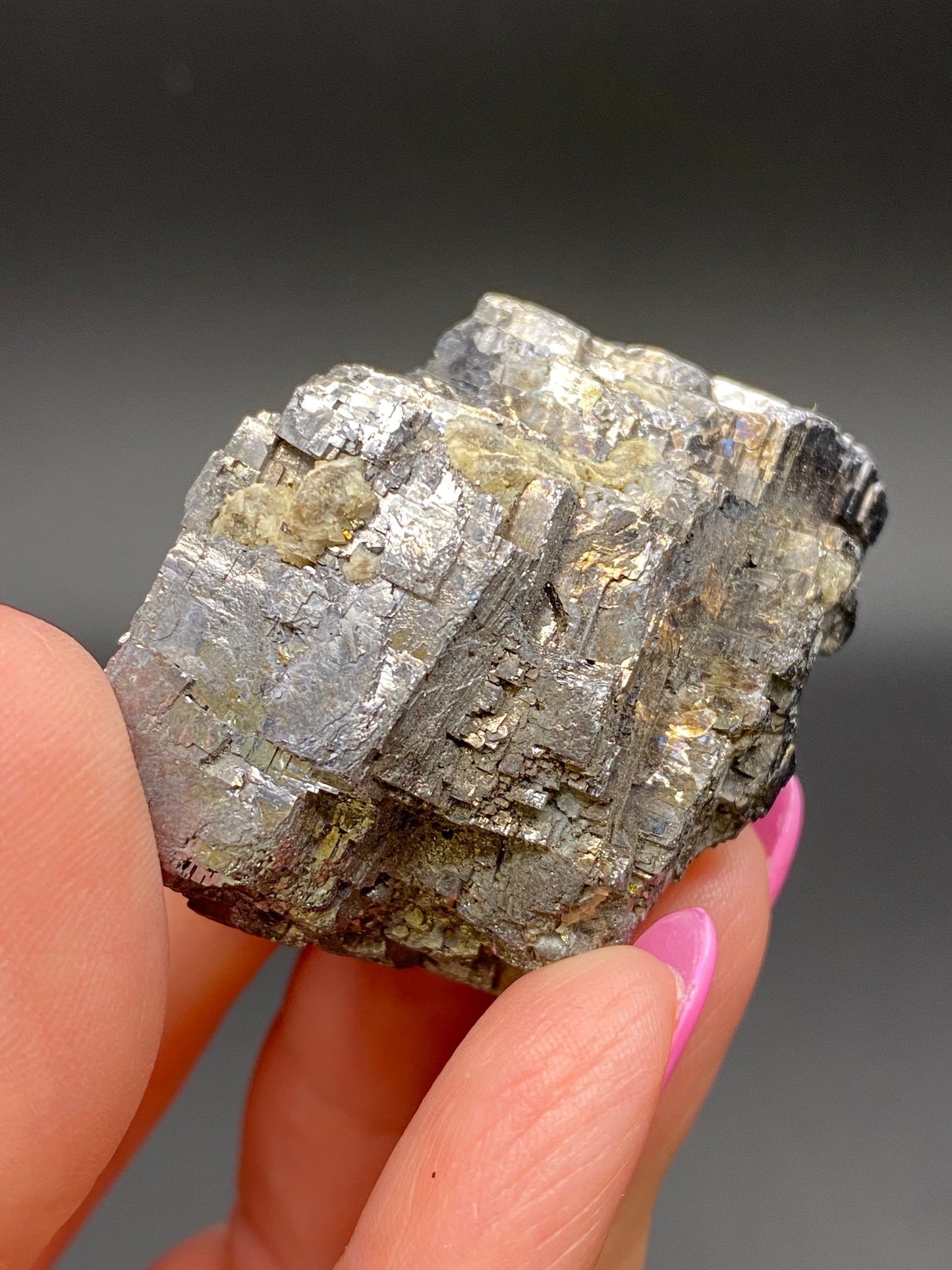 A close-up of a hand holding a metallic, reflective specimen of Galena from the Sweetwater Mine in Ellington, Reynolds County, Missouri, USA by The Crystalary against a dark background. The Galena mineral features a rough surface with various textures and shades of grey and silver. The person's nails are painted pink.