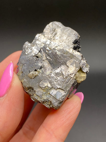 A hand with pink nail polish holds a shiny, metallic, cubic crystal. The crystal displays a silvery-gray, reflective surface and several defined geometric facets, suggesting it is a specimen of Galena from the Sweetwater Mine in Ellington, Reynolds County, Missouri. The product is known as "Galena- Sweetwater Mine, Ellington, Reynolds County, Missouri, USA" by The Crystalary. The background is dark and blurred.