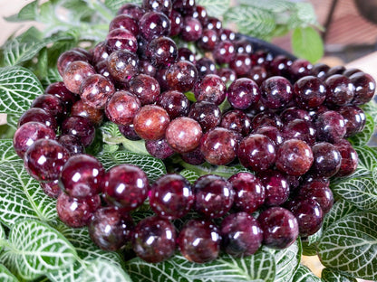 A close-up of deep purple, polished stone bead bracelets intricately coiled on a backdrop of lush, green foliage. The glossy beads are radiant, reflecting light and exhibiting variations in their rich, dark hue. These seamless designs perfectly complement The Crystalary's Garnet Crystal Bracelet- 6mm, blending harmoniously with their deep red gemstone elegance.