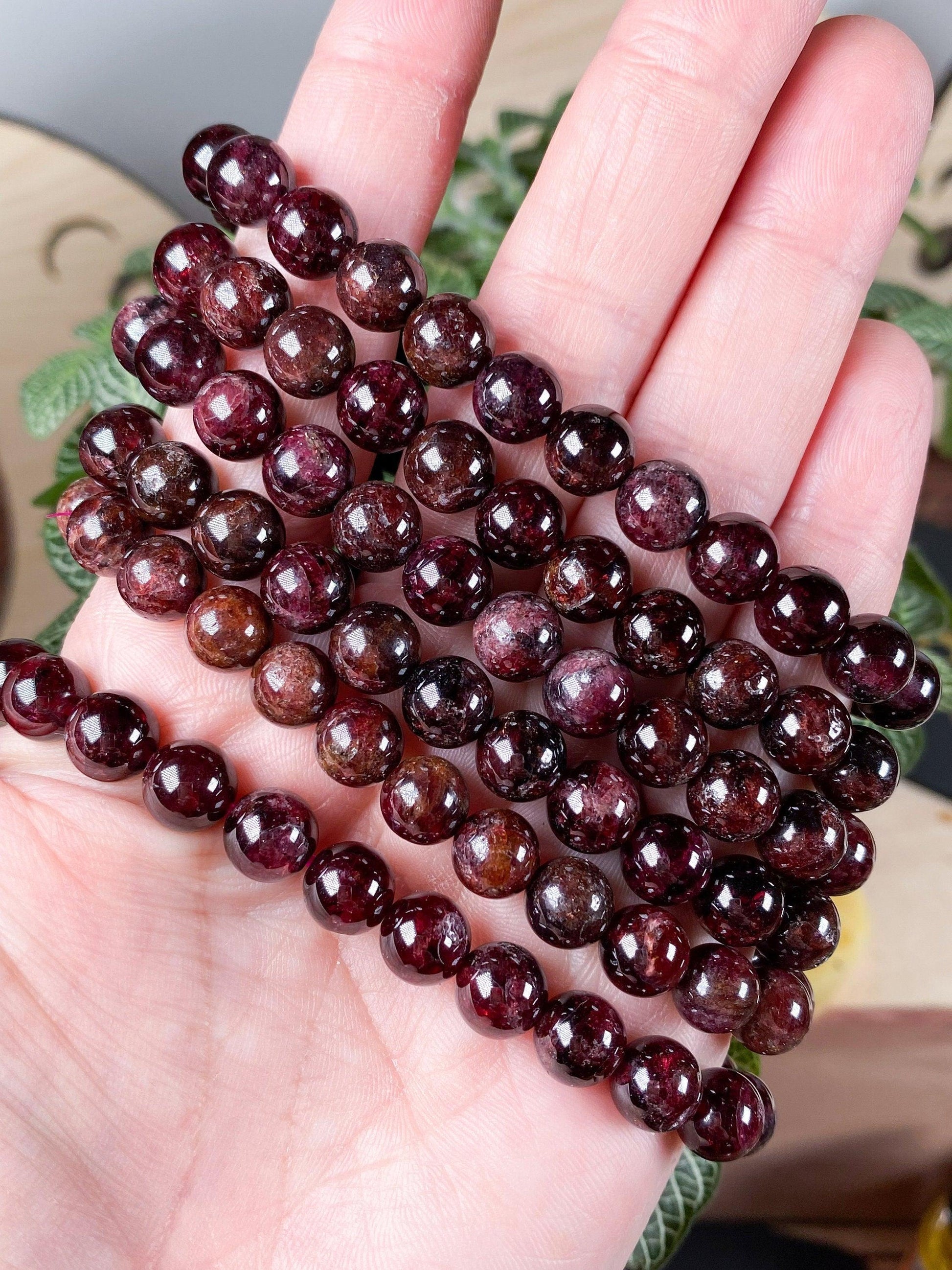 A hand holding several strands of The Crystalary's Garnet Crystal Bracelets - 6mm, showcasing varying shades of deep red and maroon polished garnet beads. The background features blurred green leaves and a wooden surface.