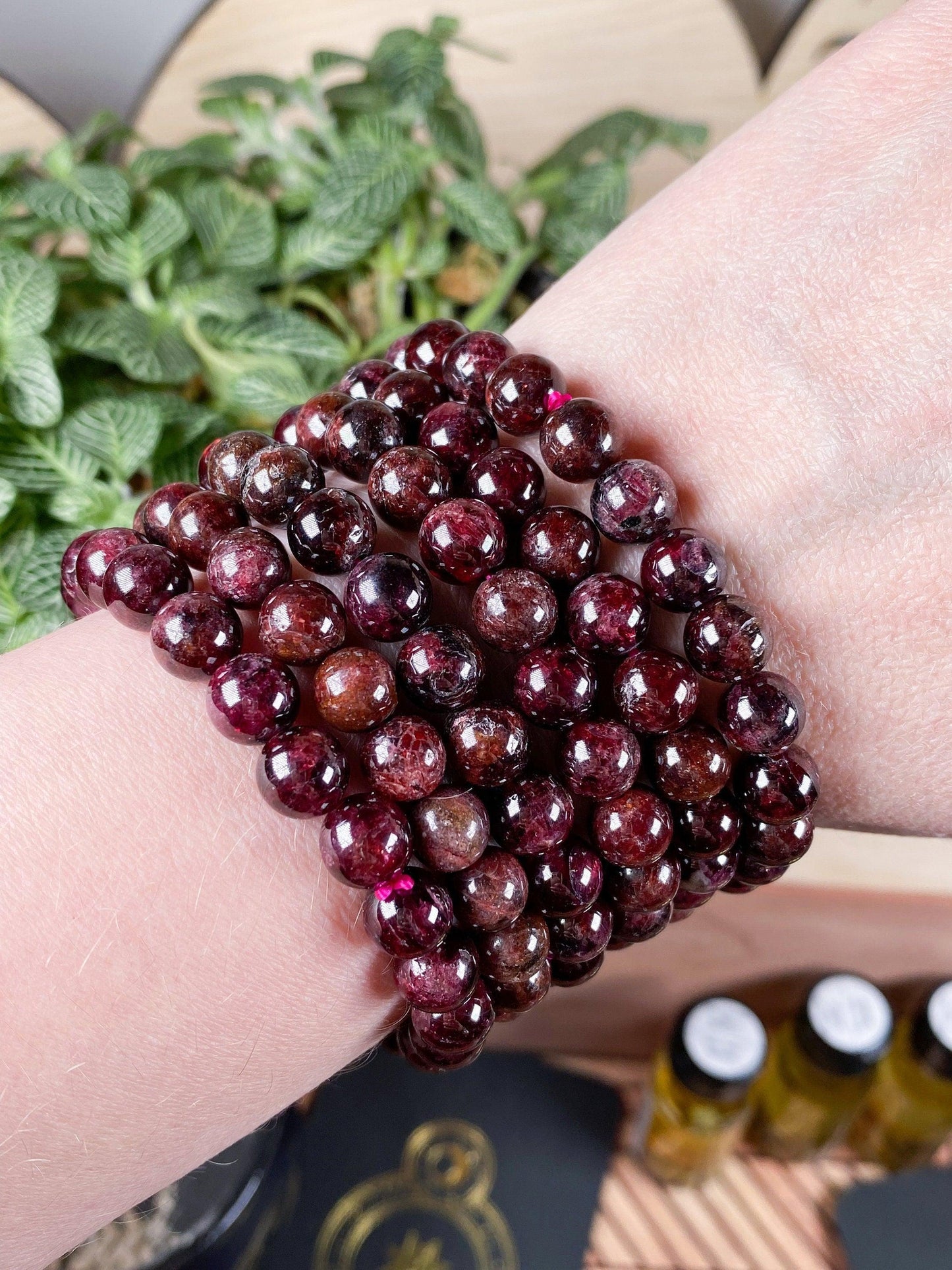 A close-up of a wrist adorned with multiple strands of The Crystalary's 6mm Garnet Crystal Bracelet. The background features leaves of a plant and small bottles of essential oils on a wooden surface.