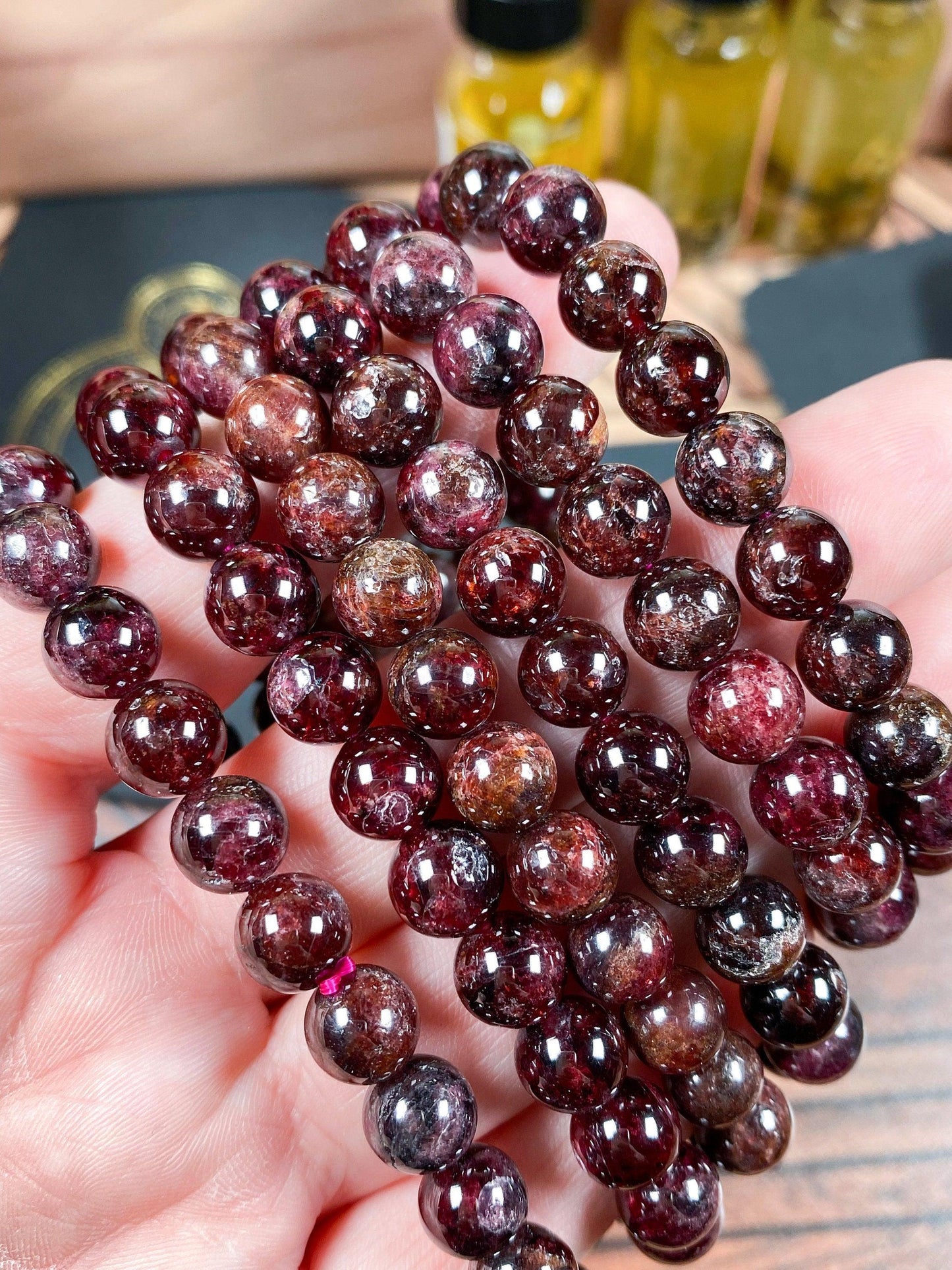 Close-up of a hand holding several strands of Garnet Crystal Bracelet-6mm beads from The Crystalary, ideal for creating stunning garnet crystal bracelets. The beads are polished and display various shades of deep red with sparkling inclusions. In the background, blurred yellow bottles and other objects add to the scene.