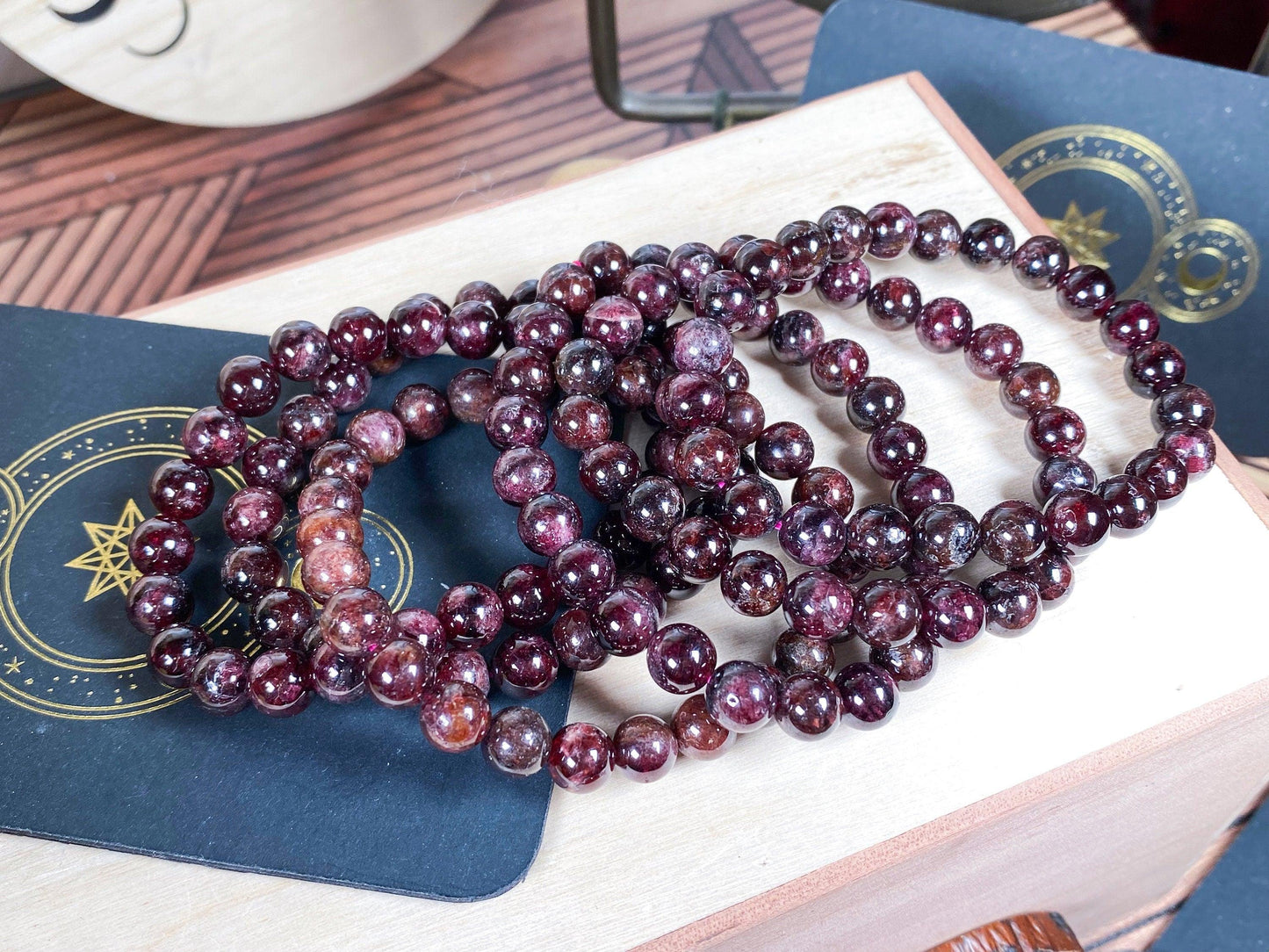 A collection of Garnet Crystal Bracelets (6mm) from The Crystalary are displayed on a wooden surface. The gemstone bracelets rest atop a couple of black cards adorned with gold geometric and celestial designs, all set against a natural wooden textured background.