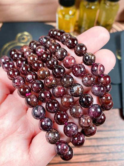 A hand holds several strands of polished, round deep red Garnet Crystal Bracelet- 6mm beads from The Crystalary, exhibiting rich hues with occasional light speckles. In the background, yellow liquid-filled bottles and a black surface with a gold design are partially visible. These Garnet Crystal Bracelets exude elegance and charm.