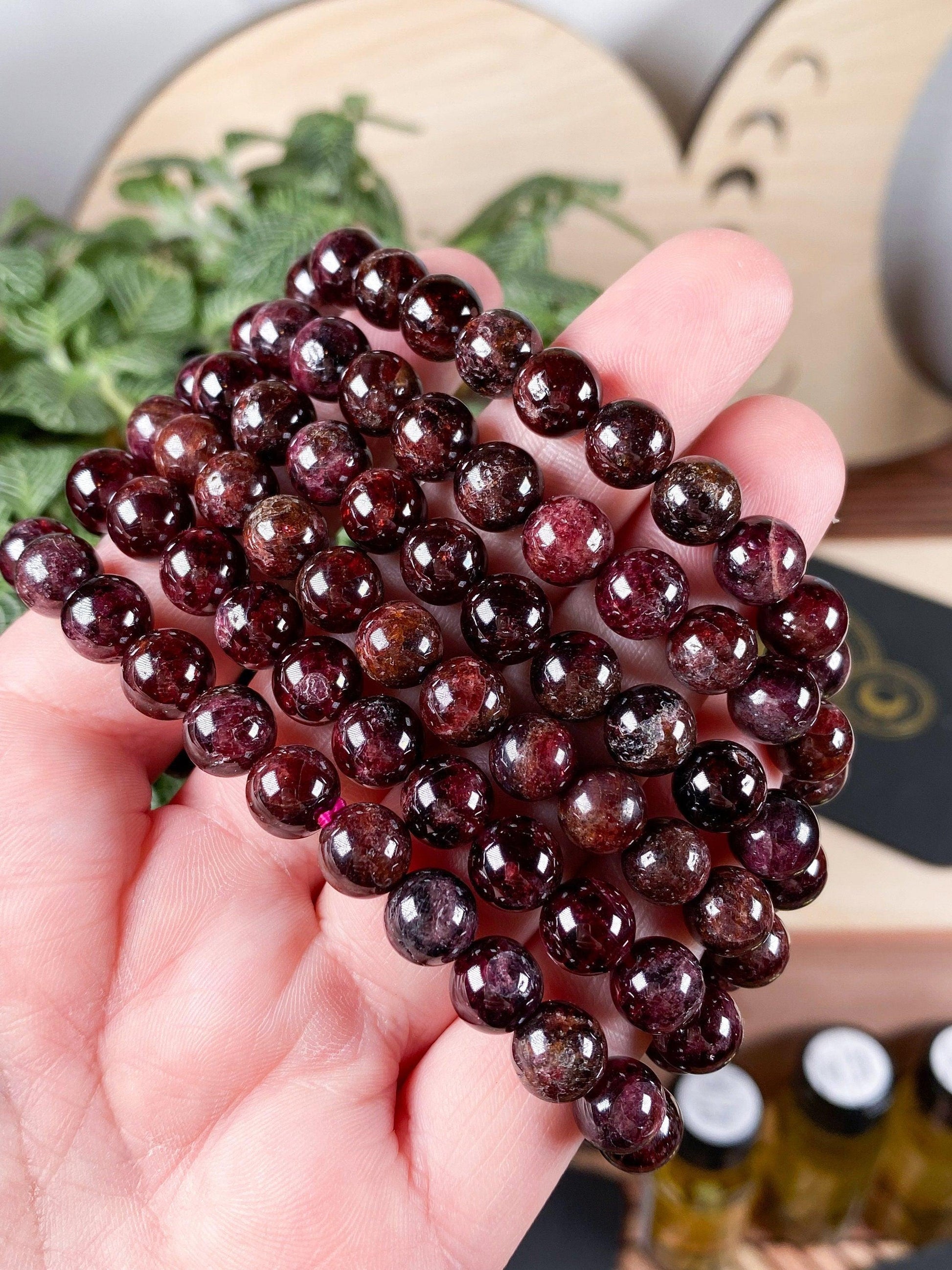 A hand holding several strands of The Crystalary's Garnet Crystal Bracelet- 6mm, each bead polished to a shiny finish. The background includes some greenery and blurred elements, creating a natural and rustic setting perfect for showcasing these garnet crystal bracelets.