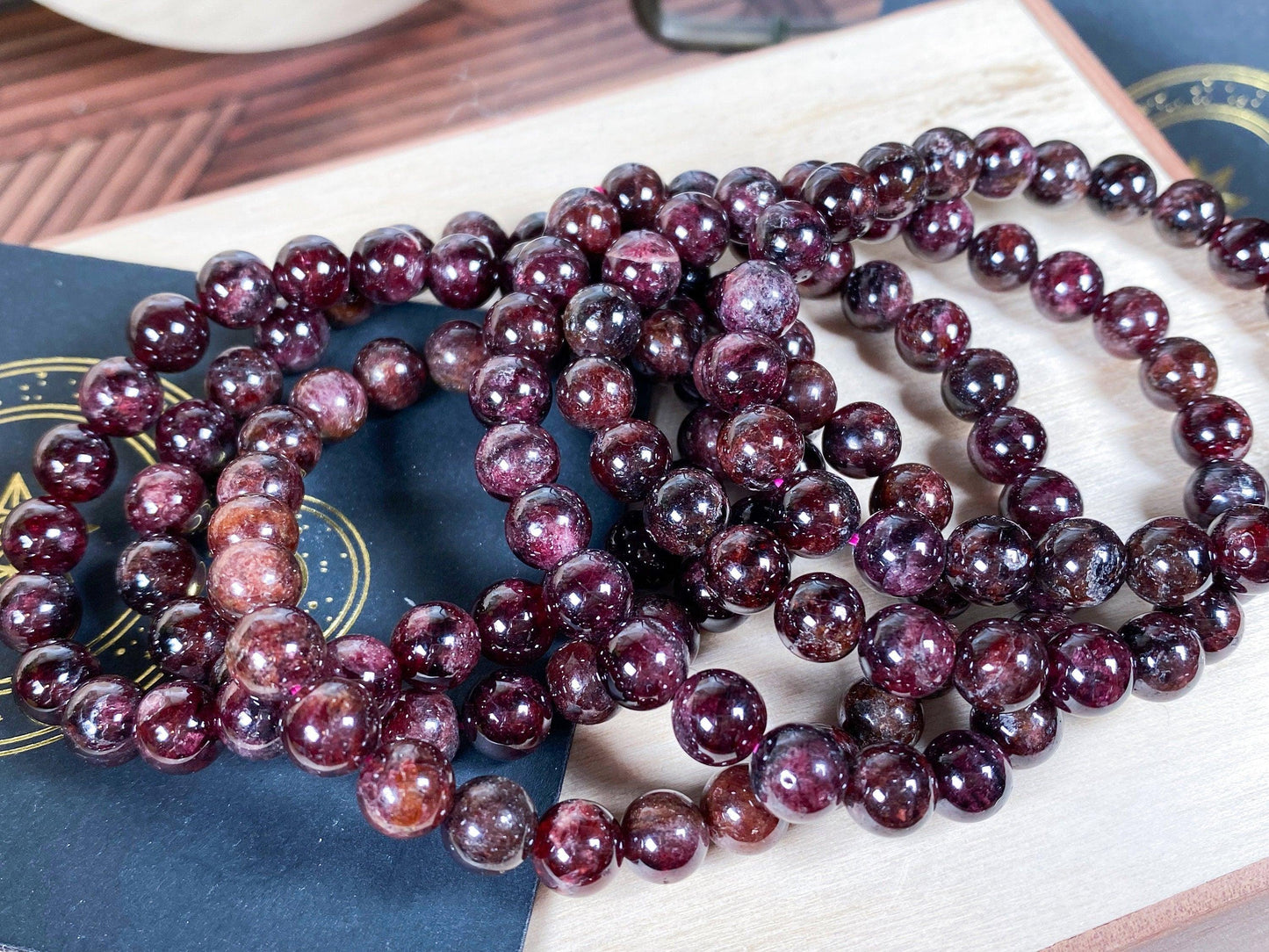 A close-up image shows several strands of deep red Garnet Crystal Bracelets- 6mm from The Crystalary, arranged neatly on a light wooden surface. The beads have a shiny, glass-like finish, reflecting the surrounding light. Parts of a dark background with golden accents are also visible.