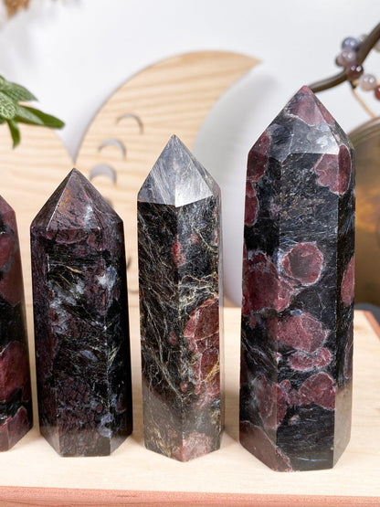 Four polished Garnet Crystal Points from The Crystalary, each featuring multicolored patterns and sharp points, are displayed on a wooden surface. The background includes wooden decorative elements and a few green leaves. These crystal points have dark bases with red and white speckles or streaks, showcasing unique colors and patterns.