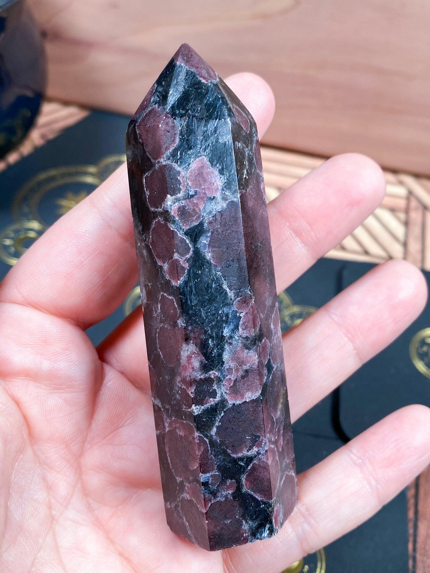 A hand holding The Crystalary's Garnet Crystal Point featuring a polished, hexagonal design with Arfvedsonite. The surface showcases irregular spots of deep red amid a black base, creating a textured and natural pattern. In the background, there's a wooden surface adorned with decorative coasters.