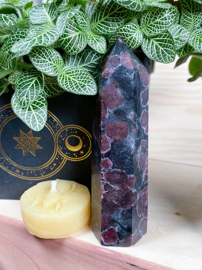 A Garnet Crystal Point from The Crystalary, featuring unique colors and patterns, stands on a wooden surface. Beside it is a small, round yellow candle carved with a floral pattern. In the background are green leafy plants and a black book with gold designs.