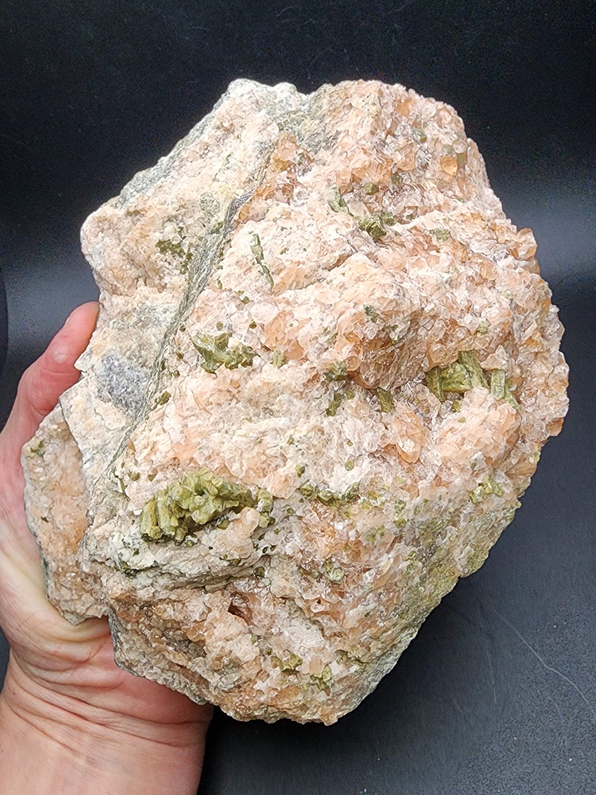 A hand holds a large, rough specimen of Garnet and Diopside from the Jeffrey Mine against a black background. The unpolished mineral, featuring a mix of pink, gray, and green crystal formations, is a stunning piece offered by The Crystalary.