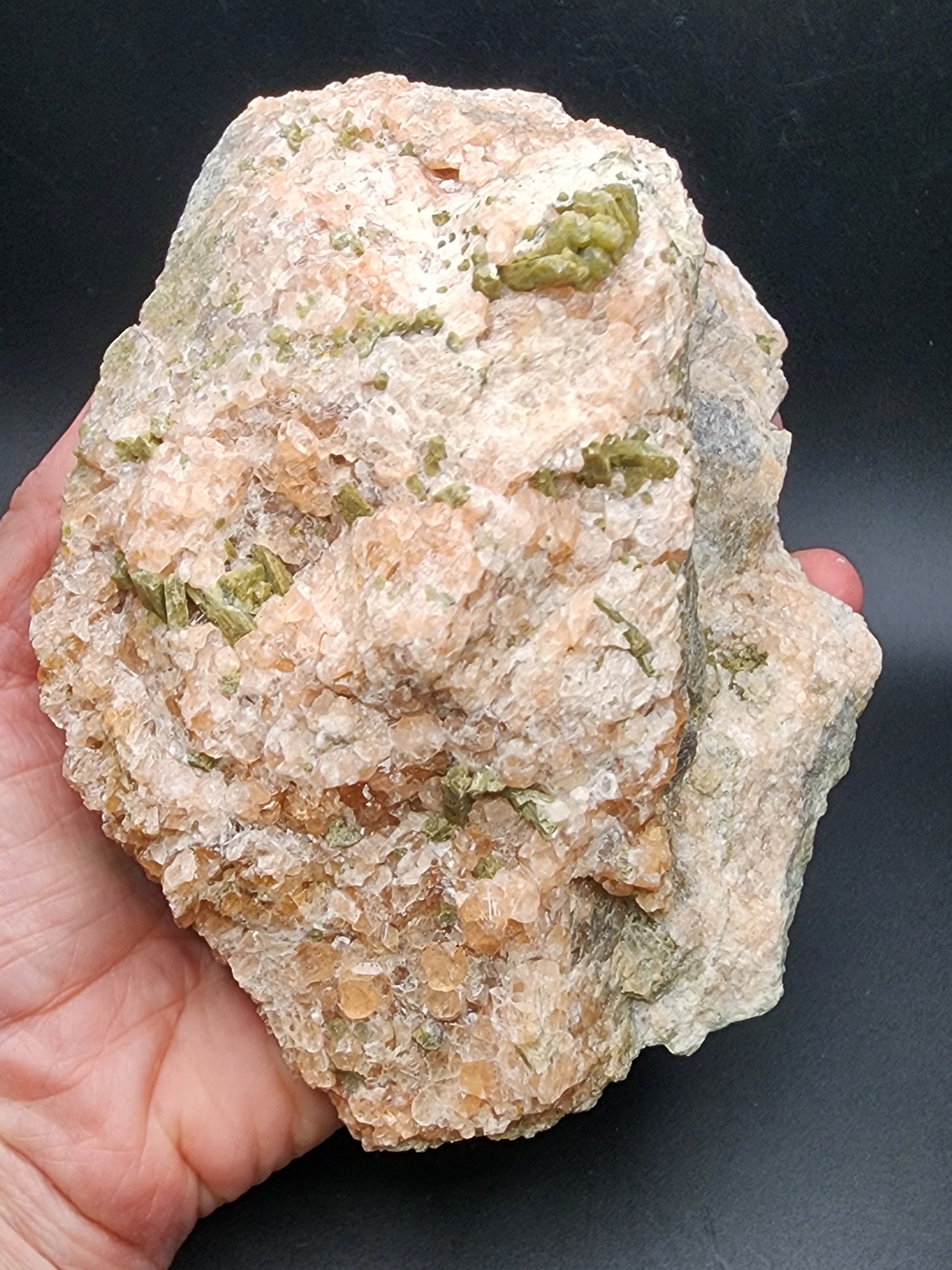 A hand holding a large rock with a rough texture. The Garnet and Diopside rock, sourced from Jeffrey Mine in Val-des-Sources, Québec, features a combination of white, pink, and green crystalline formations showcasing various minerals. The black background contrasts with the light-colored rock from The Crystalary collection, highlighting its details and textures.