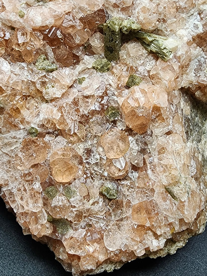 Close-up of "Garnet, Diopside" from Jeffrey Mine, Val-des-Sources, Les Sources RCM, Estrie, Québec, Canada by The Crystalary featuring clusters of translucent pale peach-pink crystals mixed with greenish diopside deposits. The crystals exhibit a shiny, faceted texture against a dark, slightly textured background.