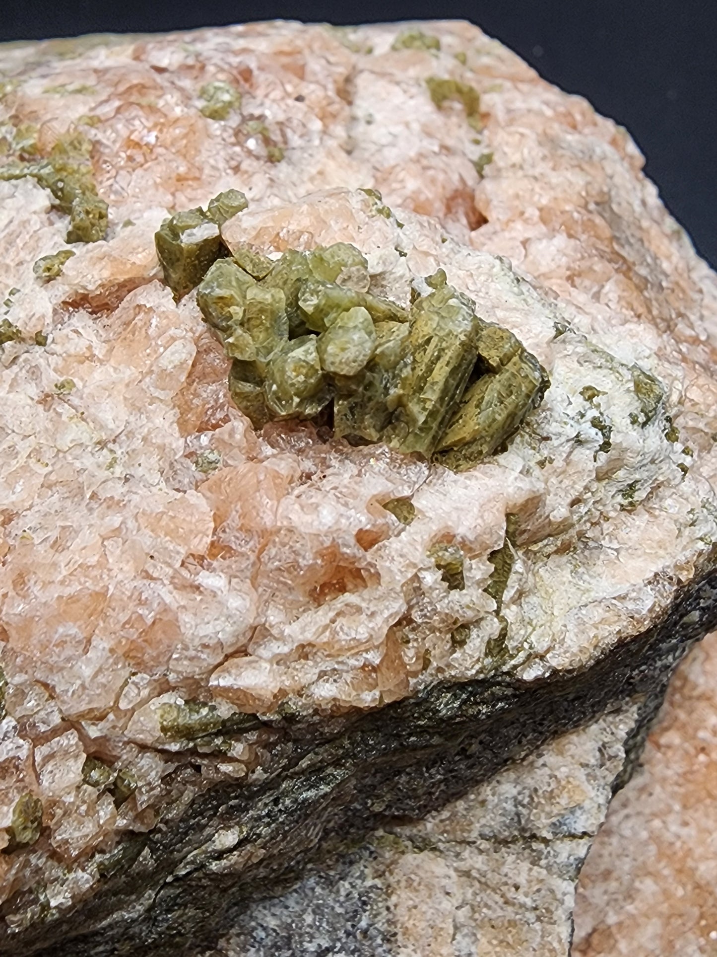 Close-up of a mineral rock featuring intricate clusters of green Diopside crystals emerging from a predominantly white and pinkish matrix. The texture of the rock is rough, highlighting the natural formation of the crystals. This specimen, known as Garnet with Diopside, hails from the renowned Jeffrey Mine in Val-des-Sources, Les Sources RCM, Estrie, Québec, Canada and is available through The Crystalary.