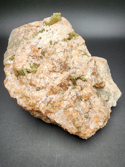 A large rock specimen from The Crystalary's collection, featuring pinkish-orange hues with small greenish Diopside crystal inclusions scattered throughout its rough surface. This stunning Garnet is reminiscent of the minerals found at Jeffrey Mine in Val-des-Sources, Les Sources RCM, Estrie, Québec, Canada. The rock sits on a dark, solid background.