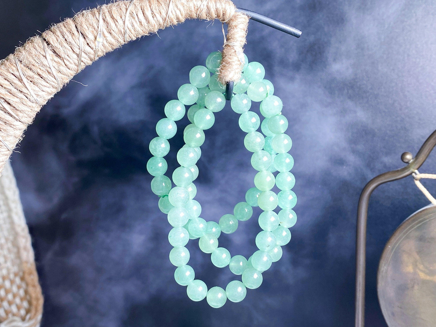 This image showcases The Crystalary's Green Aventurine Crystal Bracelet, featuring a double strand of light green beads elegantly draped over a twisted rope hanger. The smoky blue background adds a misty, ethereal quality to the scene, with an additional metallic loop partially visible on the right side.