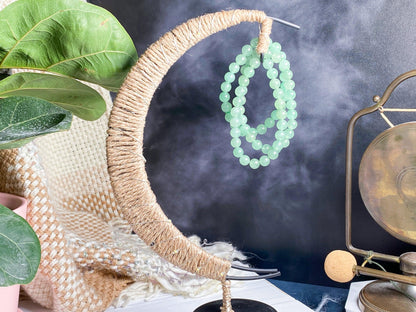 A decorative piece showcasing a crescent moon shape wrapped in twine, adorned with two of The Crystalary's Green Aventurine Crystal Bracelets in mint green, is displayed on a tabletop. Surrounding the piece are green foliage, a brass gong, and a textured blanket, creating a serene ambiance.