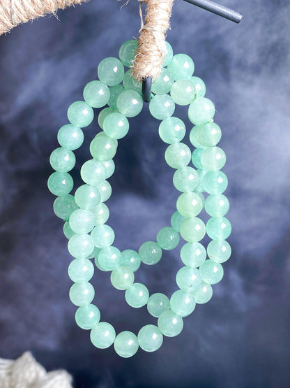 Three strands of light green jade beads are hanging from a rope, set against a blurred grayish-blue background. The beads are smooth, round, and polished, exuding an elegant appearance. The setup highlights the beads' color and texture, resembling "The Crystalary" Green Aventurine Crystal Bracelet.