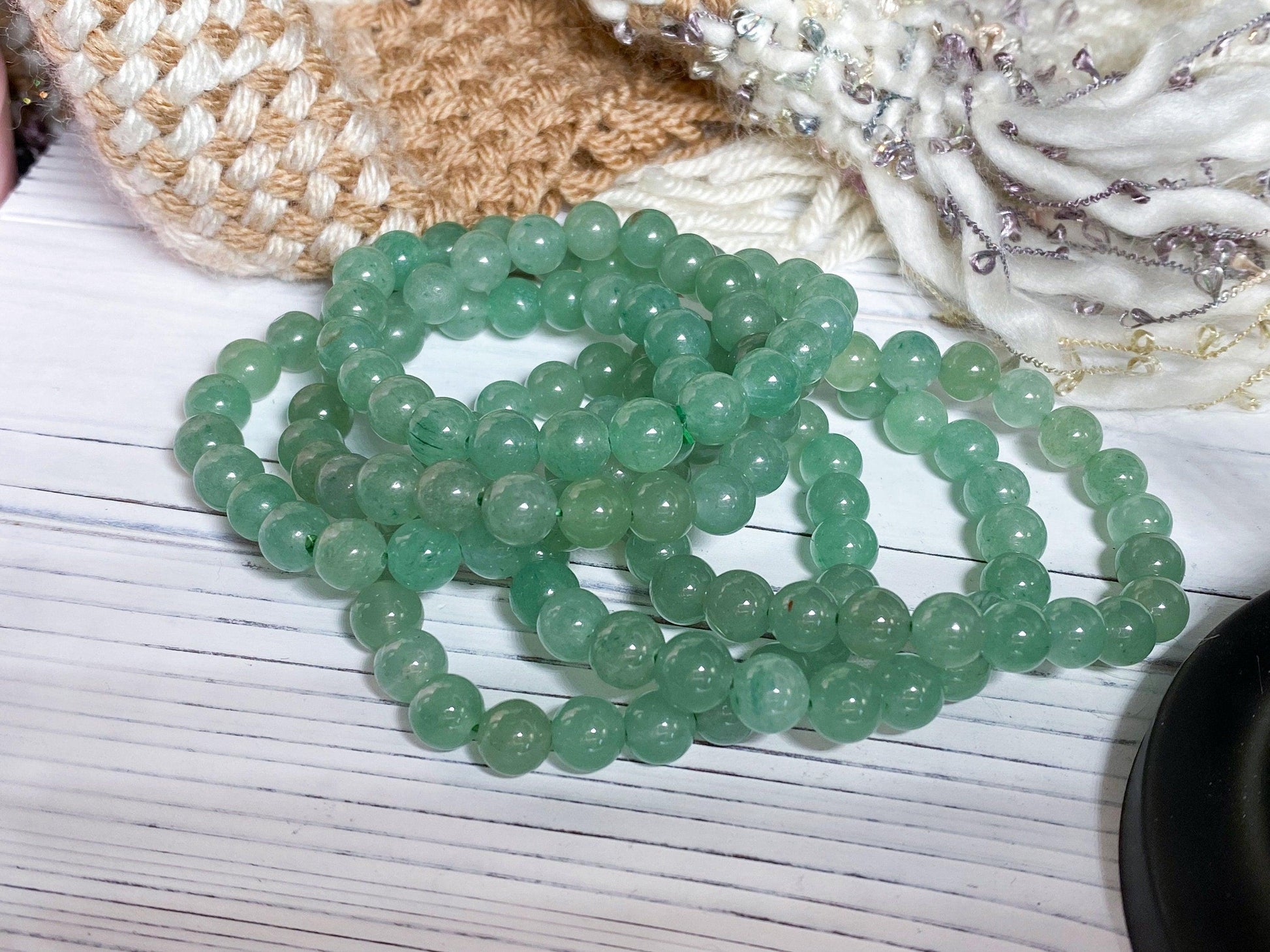 Several Green Aventurine Crystal Bracelets by The Crystalary are stacked on a white wooden surface, with textured beige and white fabrics in the background. The green beads have a polished, semi-translucent appearance, and the arrangement suggests a casual, decorative setting.