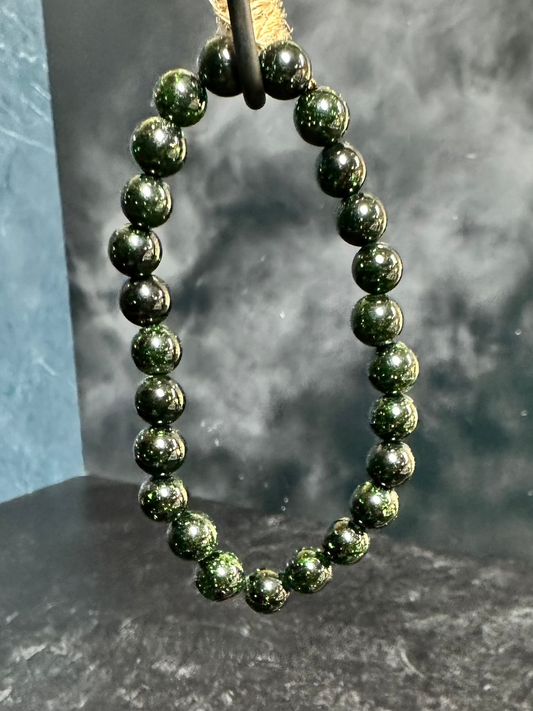 A Green Sandstone Crystal Bracelet from The Crystalary is displayed hanging from a unique curved stand wrapped in twine. The stand sits on a black speckled surface with a blue background, creating a rustic and elegant presentation. This piece joins the collection of exquisite gemstone bracelets.