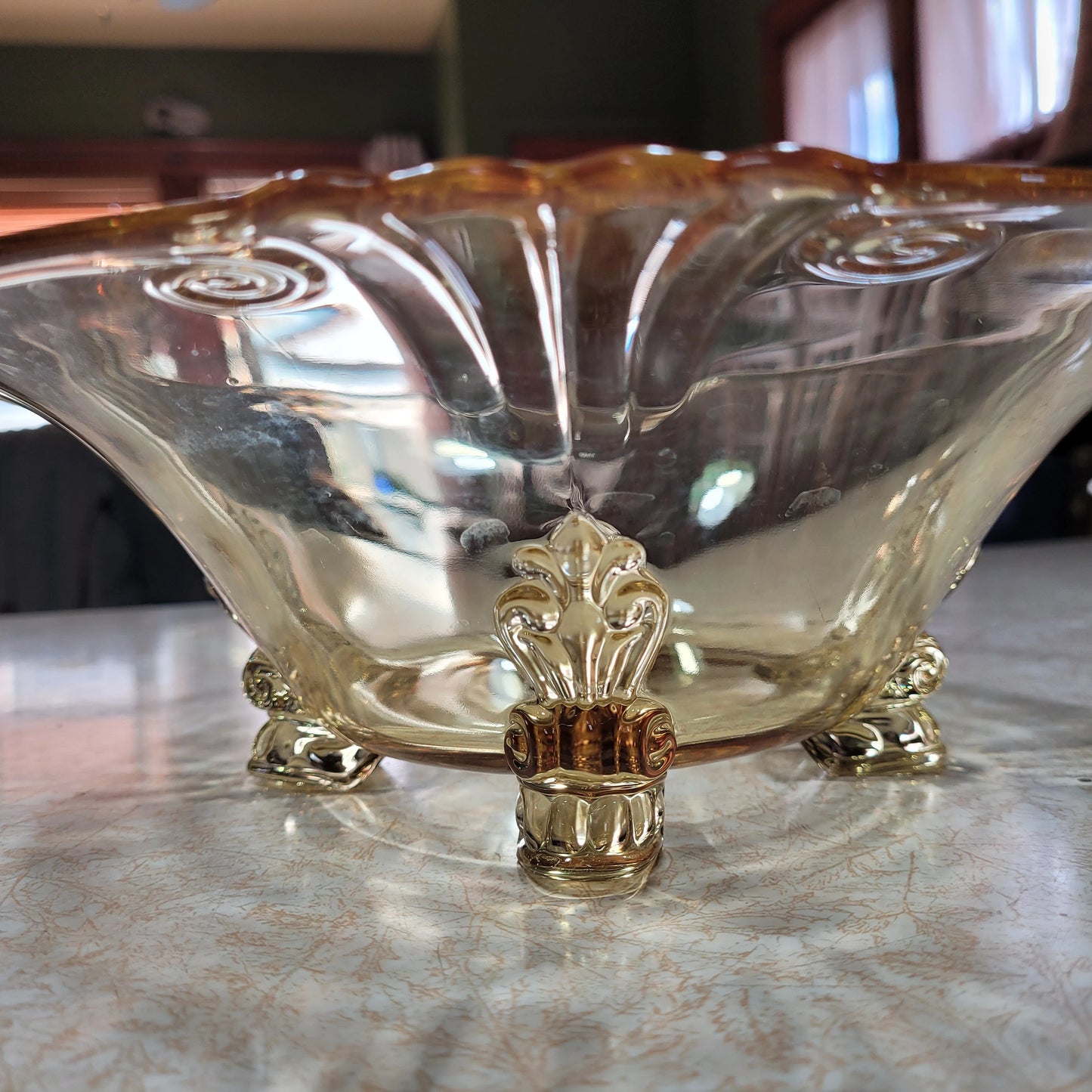 A close-up of a HEISEY Empress Sahara 4-footed dolphin foot bowl from the 1940s, resting on a countertop. The Crystalary piece, featuring a yellow tint and intricate gold-toned feet adorned with leaf and scroll motifs, exudes elegance.