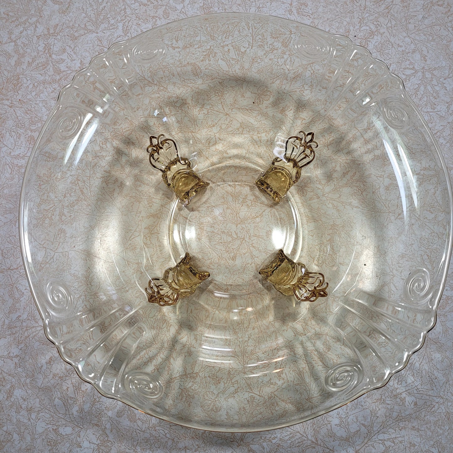 A Heisey Empress Sahara bowl with four dolphin-shaped feet, crafted in the 1940s and presented by The Crystalary, features a beautiful yellow hue. This exquisite glassware piece holds four ornate, golden-colored napkin rings, each intricately designed to resemble a crown. The rare bowl rests on a textured surface adorned with subtle floral patterns.