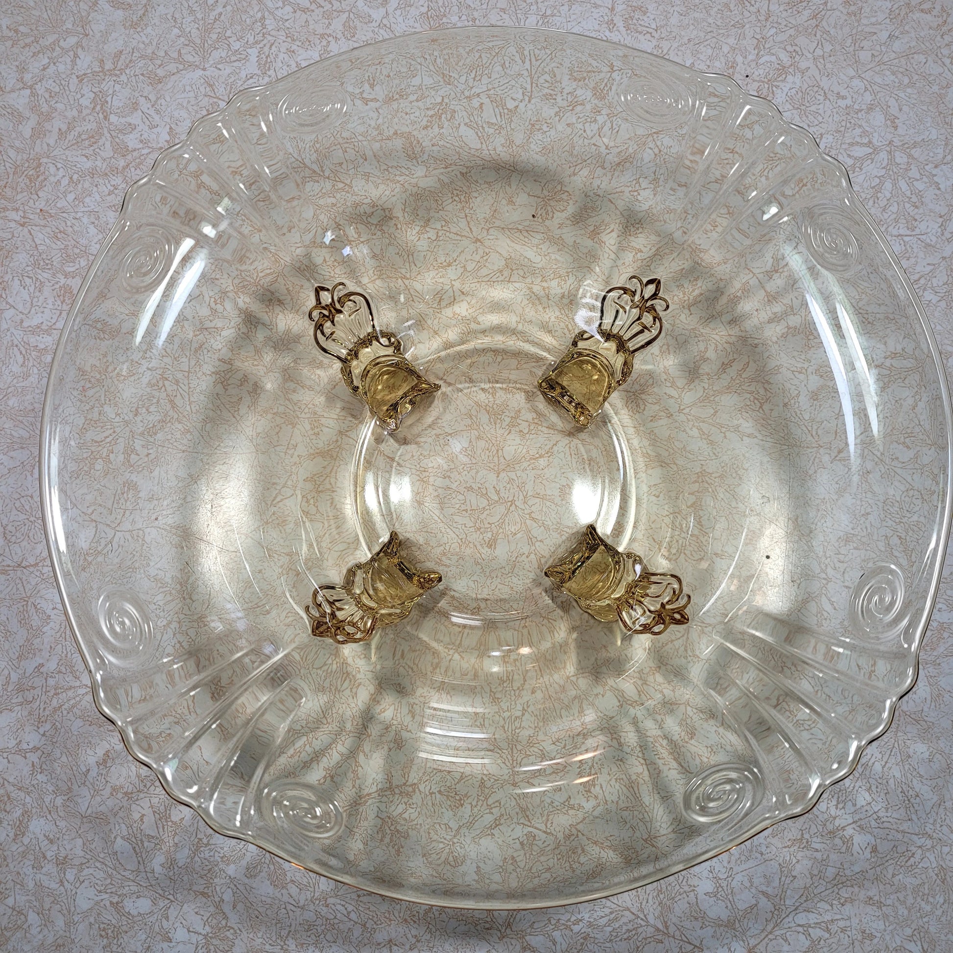 A Heisey Empress Sahara bowl with four dolphin-shaped feet, crafted in the 1940s and presented by The Crystalary, features a beautiful yellow hue. This exquisite glassware piece holds four ornate, golden-colored napkin rings, each intricately designed to resemble a crown. The rare bowl rests on a textured surface adorned with subtle floral patterns.