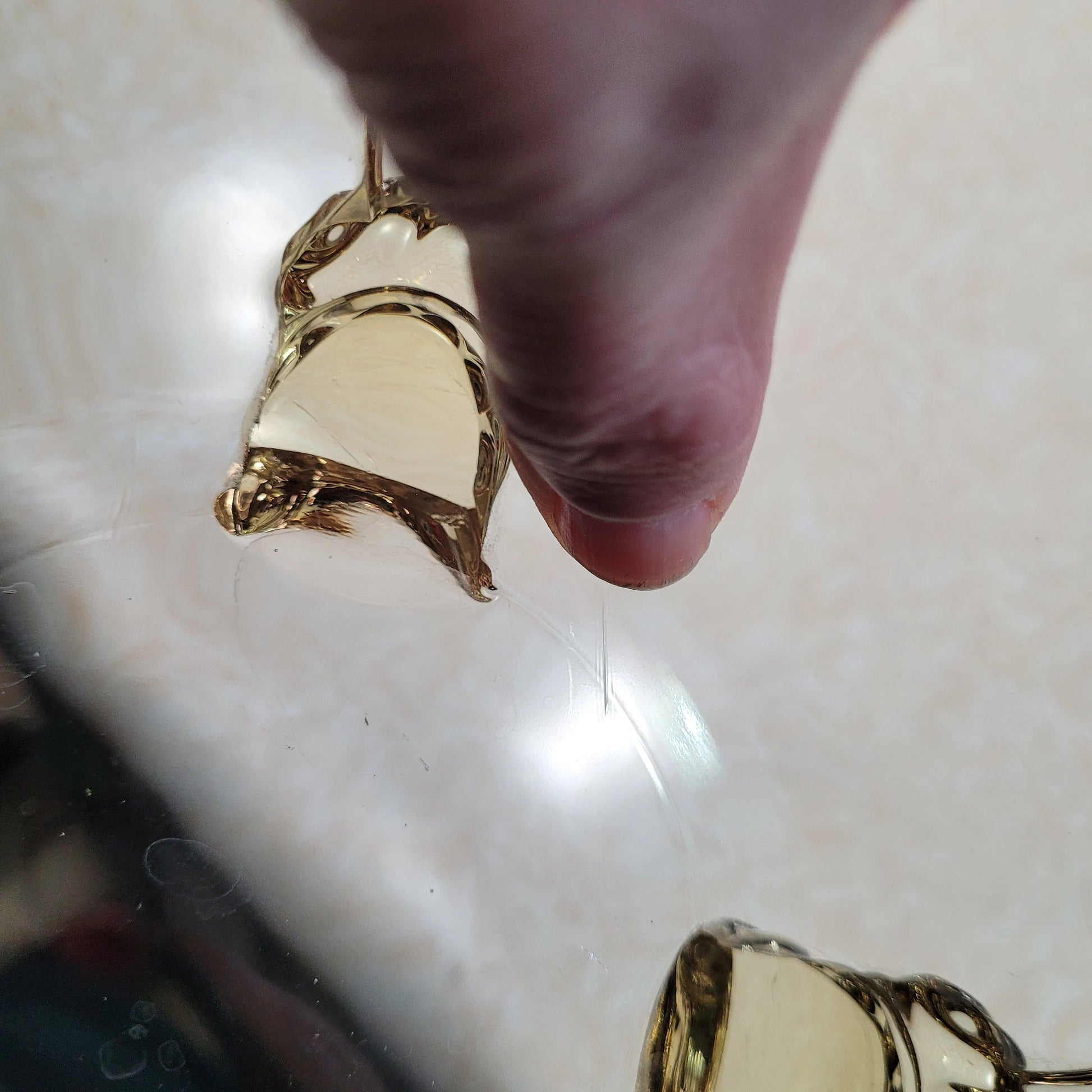 A close-up image of a person's thumb and forefinger pinching a smooth, gold-colored, transparent material reminiscent of The Crystalary's HEISEY Empress Sahara 4-footed dolphin foot bowl from the 1940s. The background is out of focus and appears to be a light surface. The material reflects light, giving it a shiny and glossy appearance.