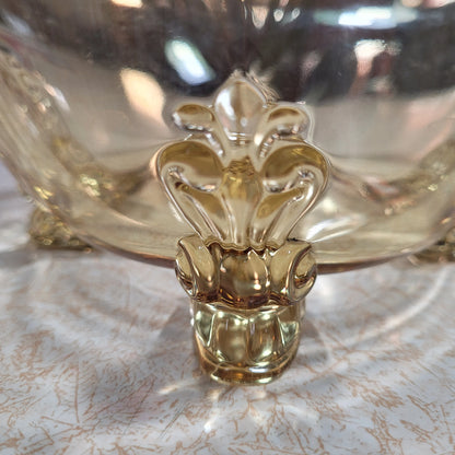 Close-up of the HEISEY Empress Sahara 4-footed dolphin foot bowl in yellow from the 1940s by The Crystalary, showcasing its ornate gold accents. The polished surface reflects light, creating a shimmering effect. The background appears blurred with a mix of warm colors and reflections.