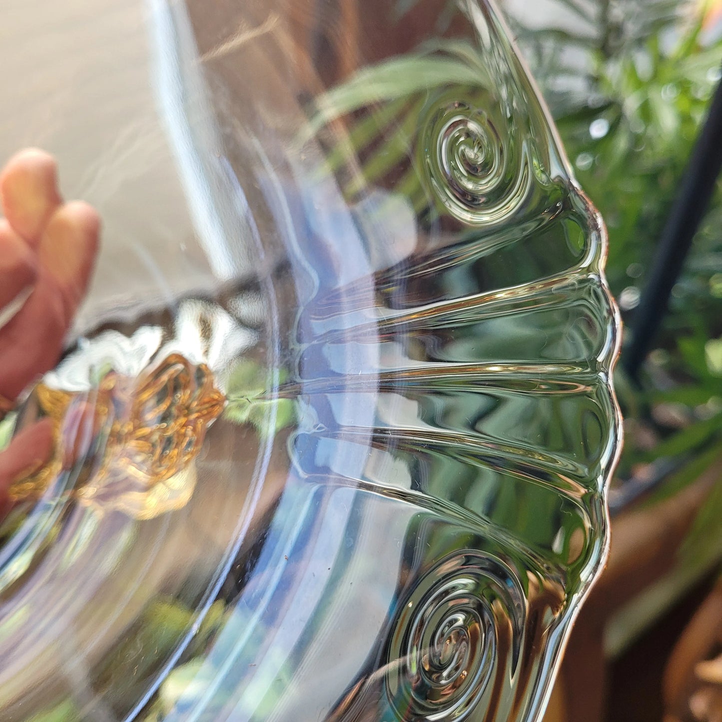 A close-up of The Crystalary's HEISEY Empress Sahara 4-footed dolphin foot bowl, held up against the light. The elegant yellow glass from the 1940s displays intricate swirling patterns and ridges along its surface. In the background, blurred green plants and part of a metal railing can be seen.