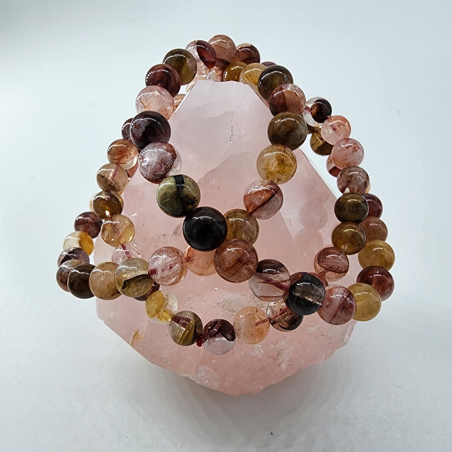 A polished pyramid-shaped rose quartz crystal from The Crystalary is elegantly paired with two Hematoid Quartz Crystal Bracelets, featuring spherical stones in pink, brown, and yellow hues. Perfectly designed for encouraging spiritual growth, the arrangement is showcased against a plain, light background.